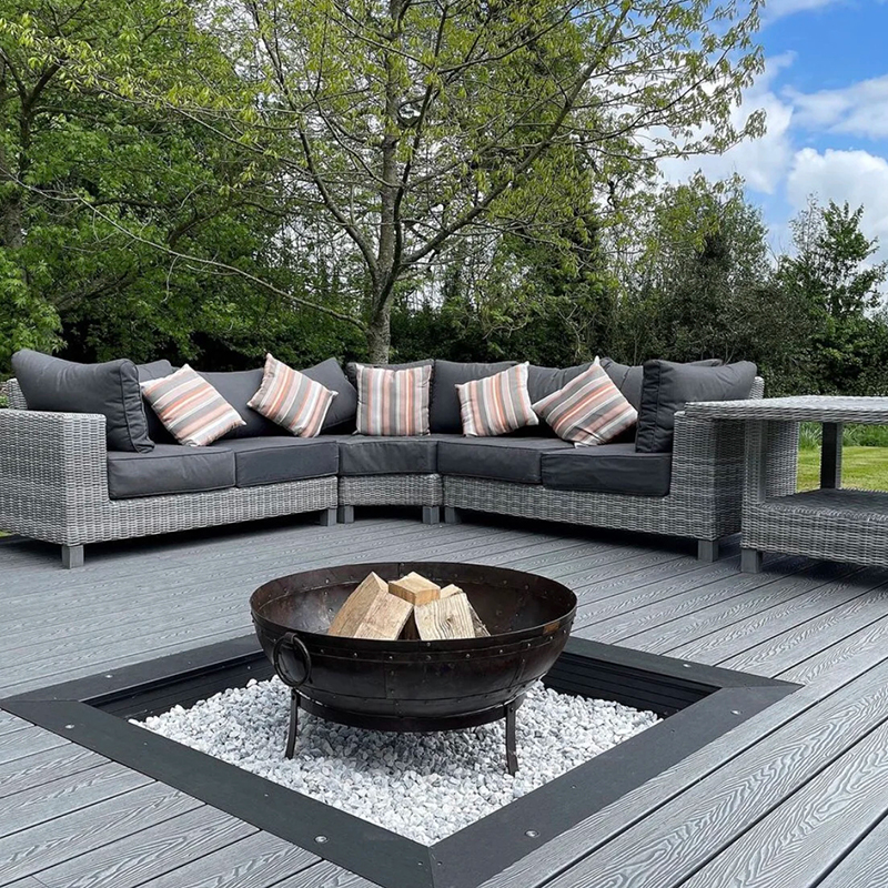Wood composite decking attached to a modern house, bordered by green bushes and small trees, with buildings and blue sky in the background.