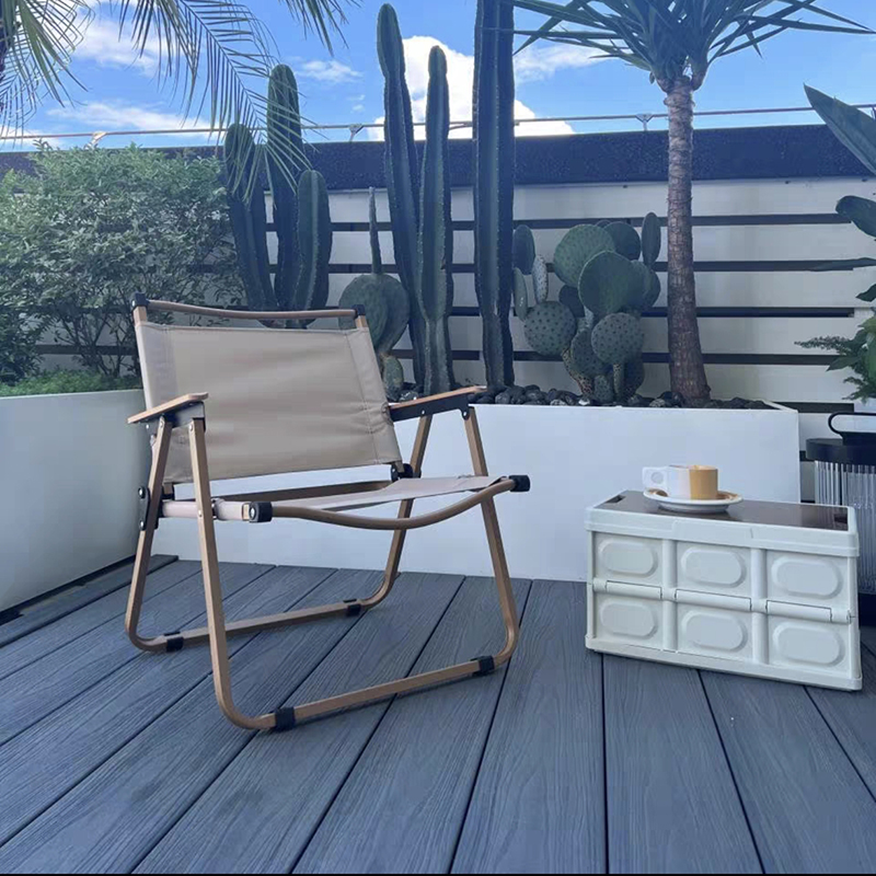 Light khaki folding chair, white Lego-style side table (with coffee set), gray wood-patterned floor. White planter holds tall cacti & palms; background has blue sky, clouds, black-white striped wall. Fresh summer vibe.
